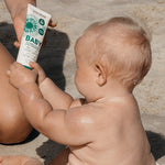 Little Urchin Baby Natural Clear Zinc Sunscreen SPF50+ 100g — an adult holding the green 100g sunscreen tube beside a baby at the beach.