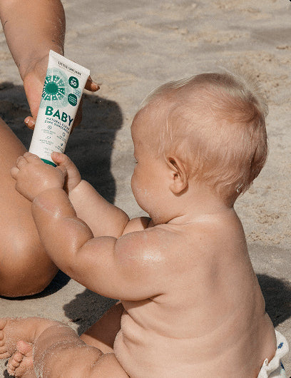 Little Urchin Baby Natural Clear Zinc Sunscreen SPF50+ 100g — an adult holding the green 100g sunscreen tube beside a baby at the beach.