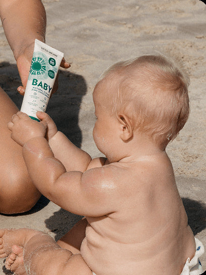 Little Urchin Baby Natural Clear Zinc Sunscreen SPF50+ 100g — an adult holding the green 100g sunscreen tube beside a baby at the beach.
