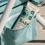 Little Urchin Baby Natural Clear Zinc Sunscreen SPF50+ 100g — the green 100g tube tucked into a striped beach bag.