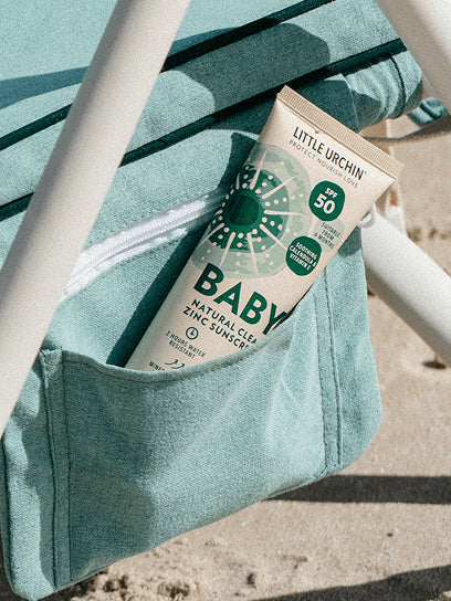Little Urchin Baby Natural Clear Zinc Sunscreen SPF50+ 100g — the green 100g tube tucked into a striped beach bag.