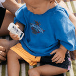 Young child seated on a picnic blanket while holding Little Urchin Baby Natural Zinc Stick Sunscreen SPF50+.