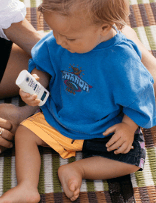 Young child seated on a picnic blanket while holding Little Urchin Baby Natural Zinc Stick Sunscreen SPF50+.