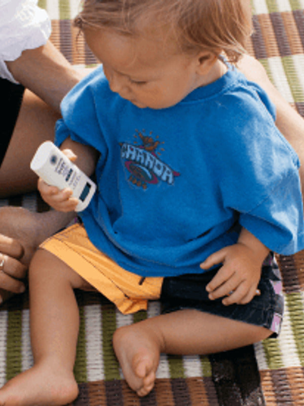 Young child seated on a picnic blanket while holding Little Urchin Baby Natural Zinc Stick Sunscreen SPF50+.
