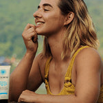 Smiling woman in a yellow swimsuit beside the water with a tube of Little Urchin Natural Clear Zinc Sunscreen SPF30 standing beside her.