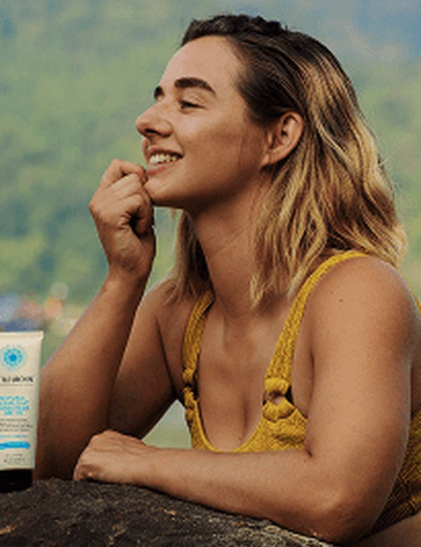Smiling woman in a yellow swimsuit beside the water with a tube of Little Urchin Natural Clear Zinc Sunscreen SPF30 standing beside her.
