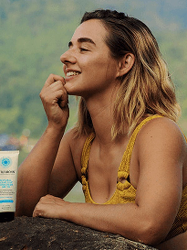 Smiling woman in a yellow swimsuit beside the water with a tube of Little Urchin Natural Clear Zinc Sunscreen SPF30 standing beside her.
