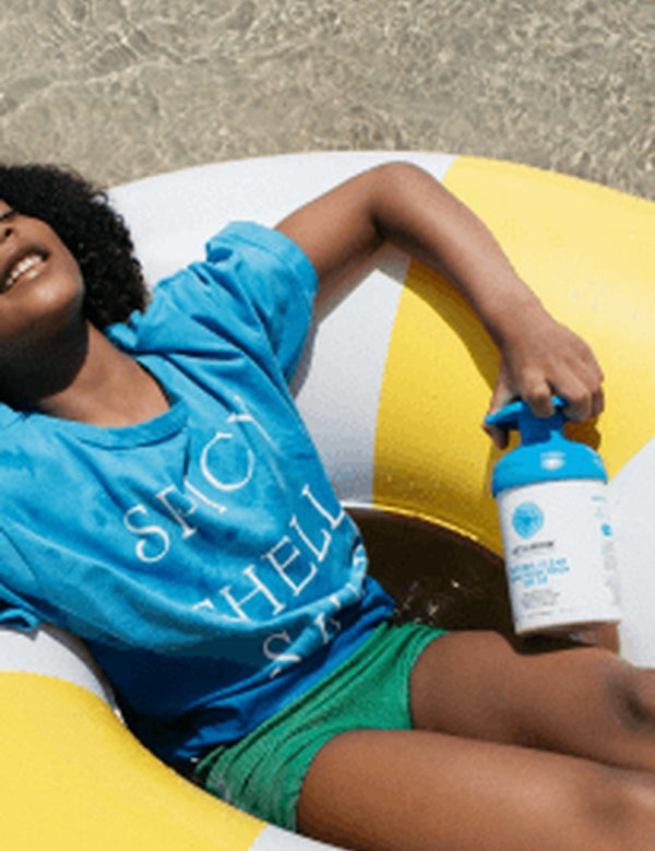 Child relaxing on a colourful inflatable ring at the beach while holding a blue-pump bottle of Little Urchin Clear Zinc Sunscreen SPF50+ 300g.