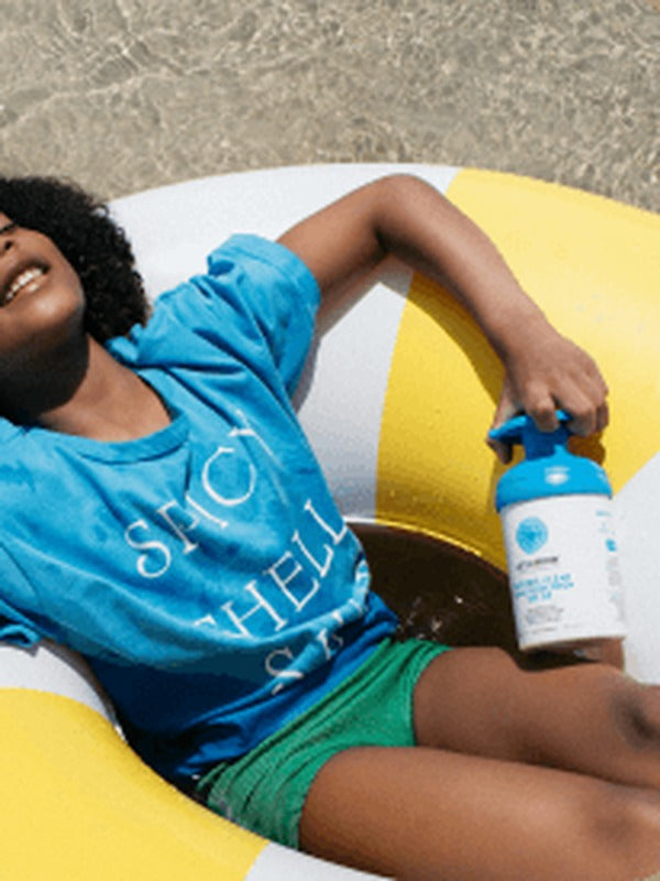 Child relaxing on a colourful inflatable ring at the beach while holding a blue-pump bottle of Little Urchin Clear Zinc Sunscreen SPF50+ 300g.