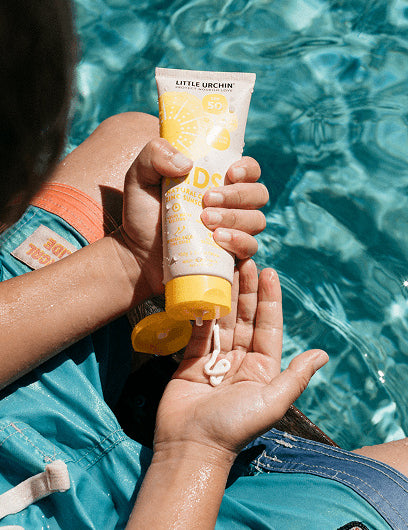 Little Urchin Kids Natural Clear Zinc Sunscreen SPF50+ 100g — hands holding the yellow 100g sunscreen tube beside a swimming pool.