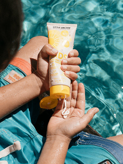 Little Urchin Kids Natural Clear Zinc Sunscreen SPF50+ 100g — hands holding the yellow 100g sunscreen tube beside a swimming pool.