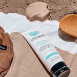 Little Urchin Natural Clear Zinc Sunscreen SPF30 100g — the 100g sunscreen tube on a beach towel beside a sun hat.