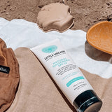 Little Urchin Natural Clear Zinc Sunscreen SPF30 100g — the 100g sunscreen tube on a beach towel beside a sun hat.