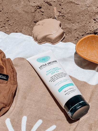 Little Urchin Natural Clear Zinc Sunscreen SPF30 100g — the 100g sunscreen tube on a beach towel beside a sun hat.
