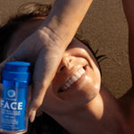 Woman lying in warm sunlight and smiling while holding a blue Little Urchin Natural Face Clear Zinc Sunscreen SPF50+ bottle beside her face.