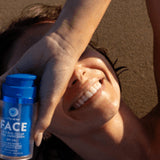 Woman lying in warm sunlight and smiling while holding a blue Little Urchin Natural Face Clear Zinc Sunscreen SPF50+ bottle beside her face.