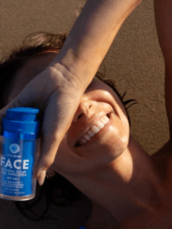 Woman lying in warm sunlight and smiling while holding a blue Little Urchin Natural Face Clear Zinc Sunscreen SPF50+ bottle beside her face.