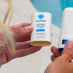 Hands holding an open Little Urchin Natural Zinc Stick Clear SPF 50+ beside its cap at a swimming pool.