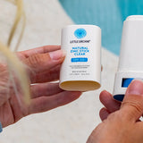 Hands holding an open Little Urchin Natural Zinc Stick Clear SPF 50+ beside its cap at a swimming pool.