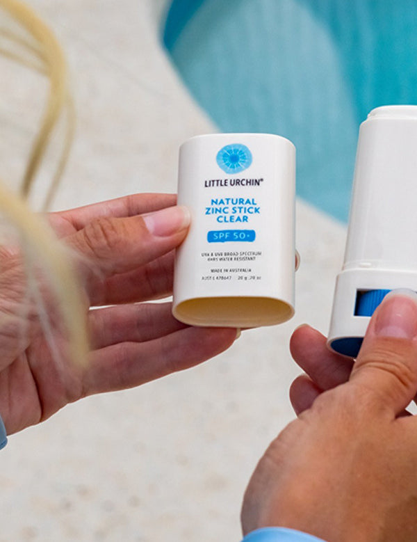 Hands holding an open Little Urchin Natural Zinc Stick Clear SPF 50+ beside its cap at a swimming pool.