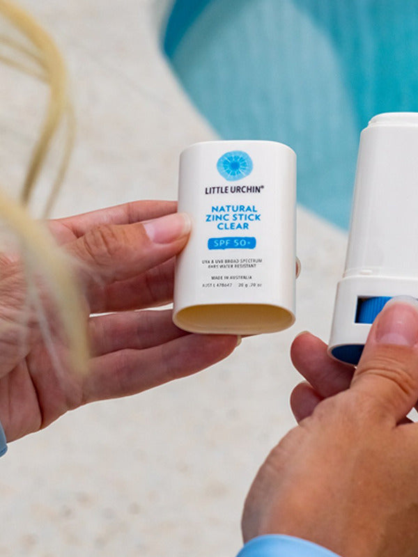 Hands holding an open Little Urchin Natural Zinc Stick Clear SPF 50+ beside its cap at a swimming pool.