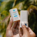 Little Urchin Natural Zinc Stick Clear SPF 50+ 20g — hands holding the clear zinc stick and its cap beneath palm trees.