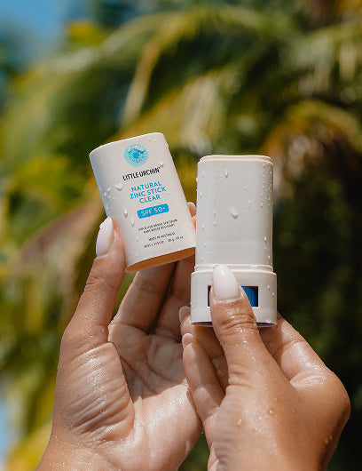 Little Urchin Natural Zinc Stick Clear SPF 50+ 20g — hands holding the clear zinc stick and its cap beneath palm trees.