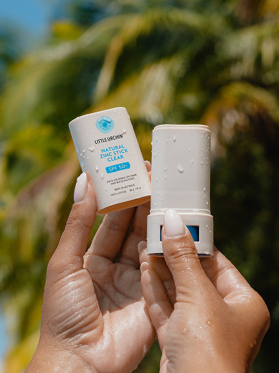 Little Urchin Natural Zinc Stick Clear SPF 50+ 20g — hands holding the clear zinc stick and its cap beneath palm trees.