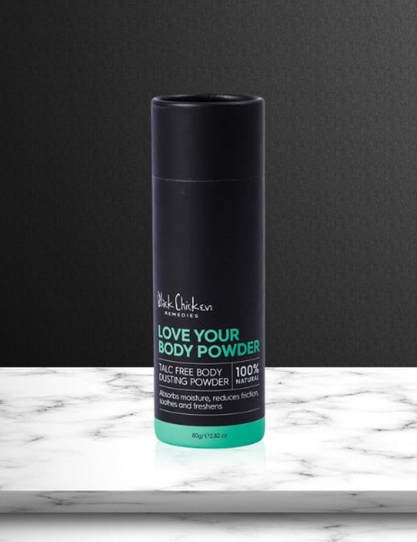 Tall black Love Your Body Powder cylinder with green lettering and a green base band standing on a white marble slab against a dark wall.