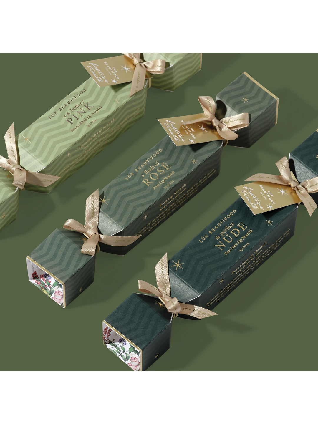 Three Luk Beautifood Christmas lip crackers in light and dark green packaging, arranged diagonally on a green background with gold ribbon bows and gift tags.