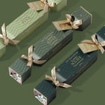 Three Luk Beautifood Christmas lip crackers in light and dark green packaging, arranged diagonally on a green background with gold ribbon bows and gift tags.