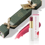 A Flush of Rose Bon Bon: dark green Christmas cracker with gold ribbons, Rosé Lip Nourish lipstick, its white carton and a wine-red colour smear.