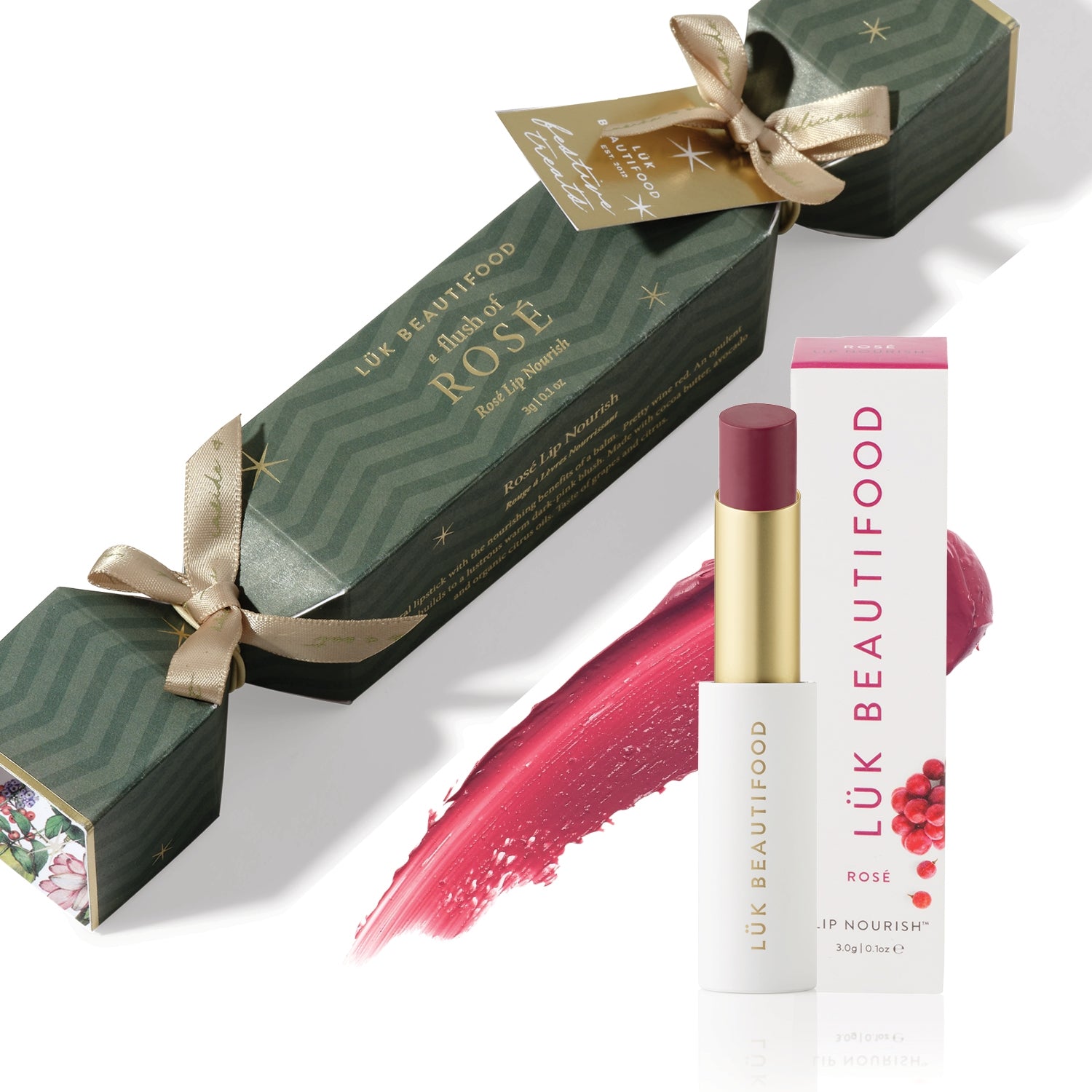 A Flush of Rose Bon Bon: dark green Christmas cracker with gold ribbons, Rosé Lip Nourish lipstick, its white carton and a wine-red colour smear.
