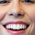 Close-up of a woman smiling broadly, showing her teeth and softly glossy pink lips wearing Luk Beautifood Lip Nourish lipstick in Nude Pink.