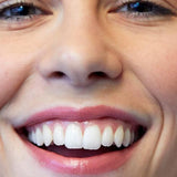 Close-up of a woman smiling broadly, showing her teeth and softly glossy pink lips wearing Luk Beautifood Lip Nourish lipstick in Nude Pink.