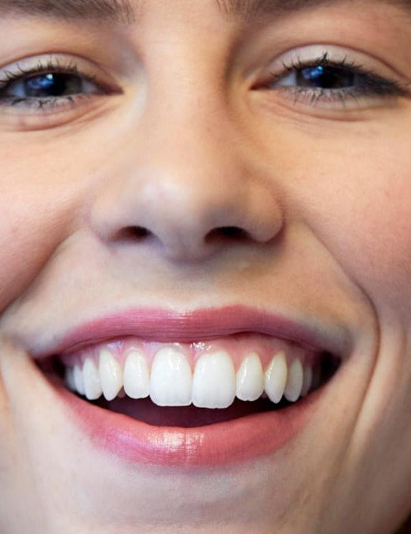 Close-up of a woman smiling broadly, showing her teeth and softly glossy pink lips wearing Luk Beautifood Lip Nourish lipstick in Nude Pink.