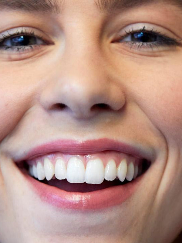 Close-up of a woman smiling broadly, showing her teeth and softly glossy pink lips wearing Luk Beautifood Lip Nourish lipstick in Nude Pink.