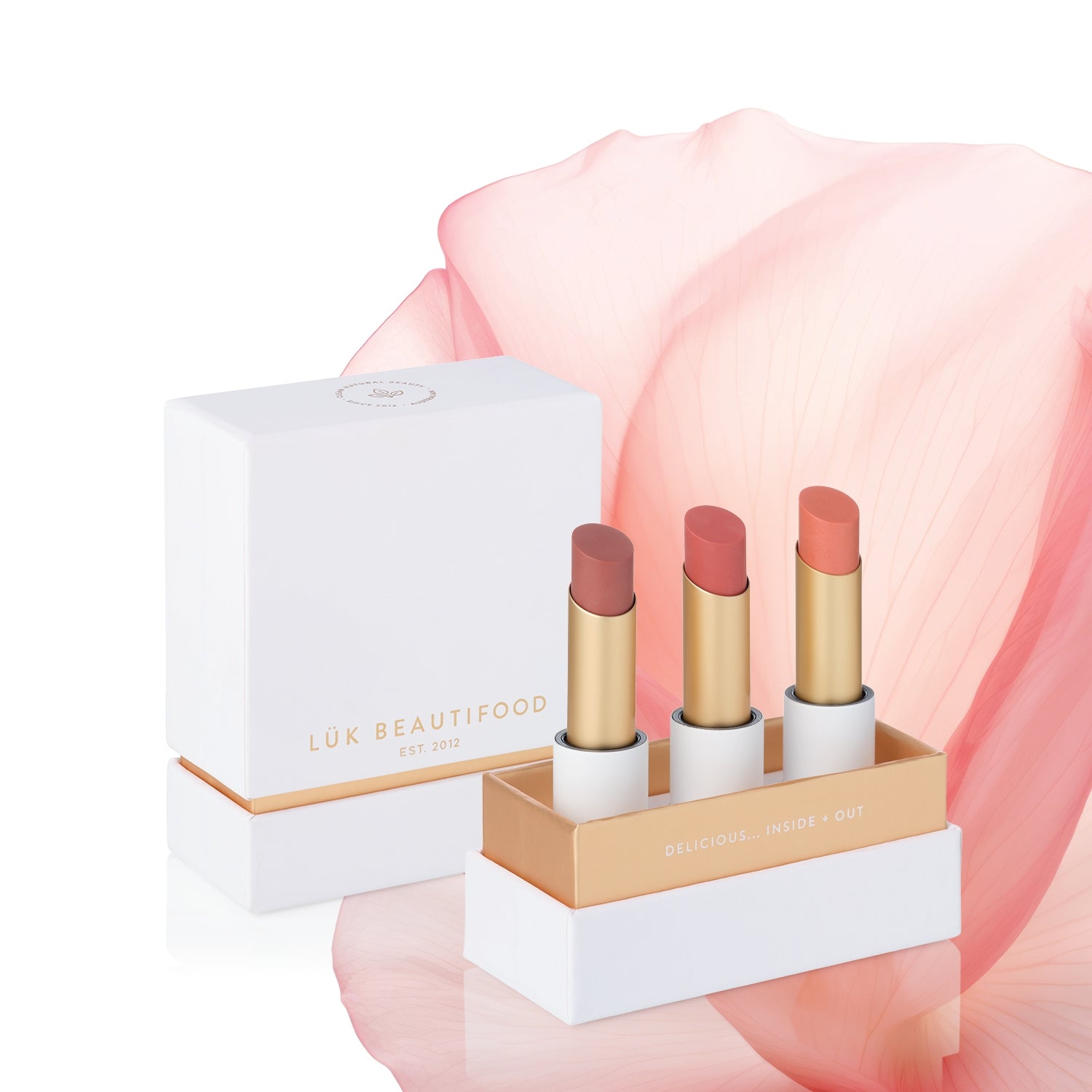 Perfect Nudes Lip Nourish Trio with Tea Rose, Nude Pink and Peach Melon lipsticks in an open white and gold gift box, with its lid and a large pink petal behind.