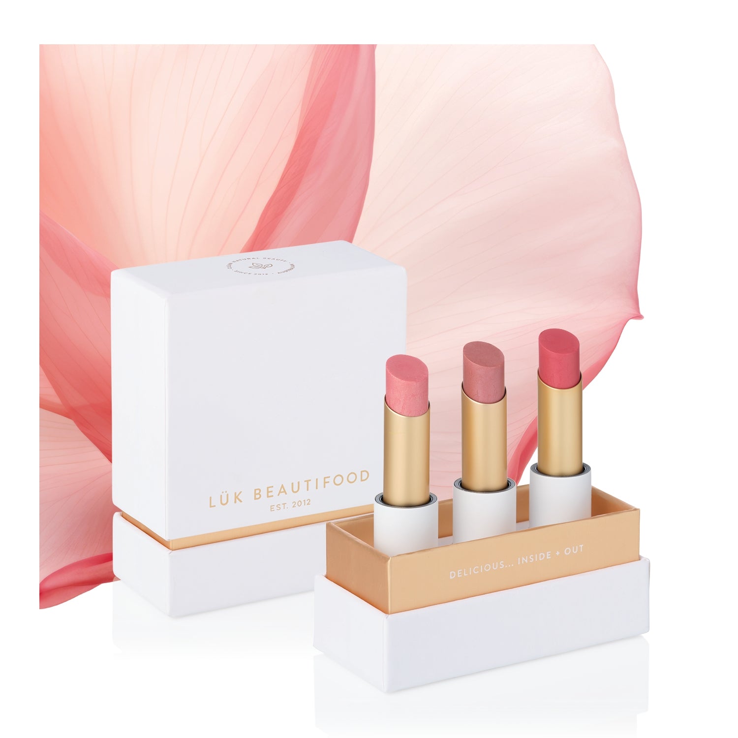 Buttery Pinks Lip Nourish Trio: Guava Blush, Pink Juniper and Ruby Grapefruit lipsticks in a white and gold presentation box beside its lid and a pink petal.