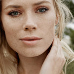 Outdoor portrait of a blonde woman with visible freckles and naturally hydrated skin, resting one hand against her neck.