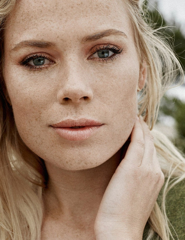 Outdoor portrait of a blonde woman with visible freckles and naturally hydrated skin, resting one hand against her neck.