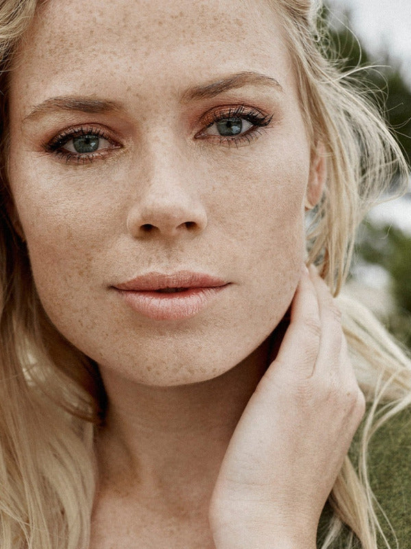 Outdoor portrait of a blonde woman with visible freckles and naturally hydrated skin, resting one hand against her neck.