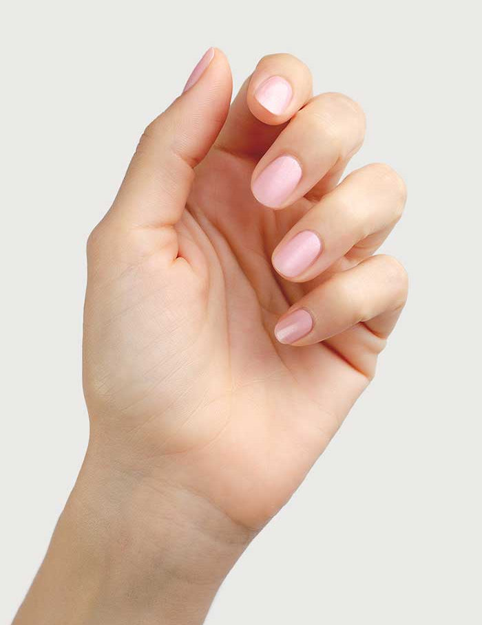 A hand with neatly manicured nails painted in Sienna Byron Bay Nail Polish - Magic, a soft, pale cherry blossom pink shade, is held up against a plain light background.