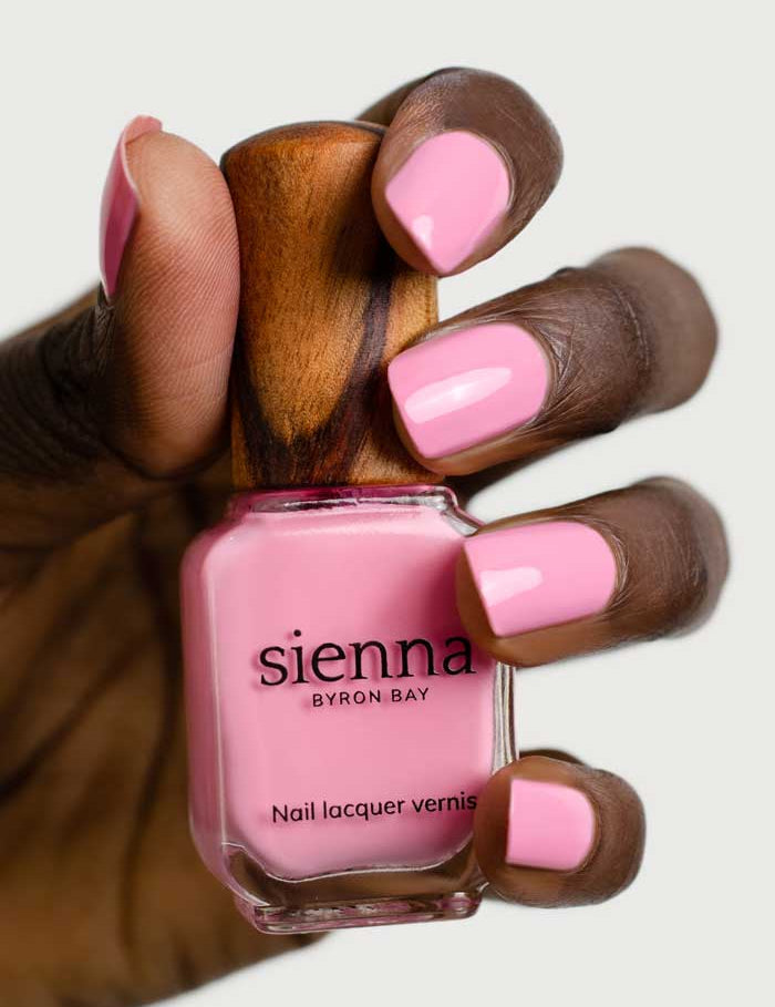 A hand with dark skin holds a bottle of Sienna Byron Bay Nail Polish - Magnolia, a plant-derived, toxin-free polish in light pink. The manicured nails are painted to match the soft Magnolia shade in the bottle.