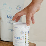 Hand resting on a white Man21 tub on a small wooden stool, with a large matching refill pouch standing behind it.