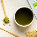 Matcha Maiden bamboo chasen whisk styled beside prepared matcha, matcha powder and bamboo tools.