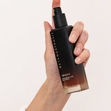 Hand holding the tall black-to-amber Metamorphosis Awaken Hyaluronic Acid Prep Spritz bottle upright with its spray pump exposed.