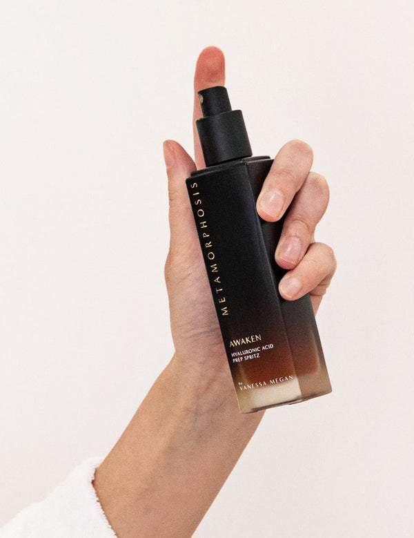 Hand holding the tall black-to-amber Metamorphosis Awaken Hyaluronic Acid Prep Spritz bottle upright with its spray pump exposed.