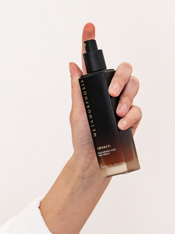 Hand holding the tall black-to-amber Metamorphosis Awaken Hyaluronic Acid Prep Spritz bottle upright with its spray pump exposed.