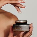 A person holding an open Metamorphosis Emerge cream jar in one hand while lifting a small dab of cream from it on a fingertip.